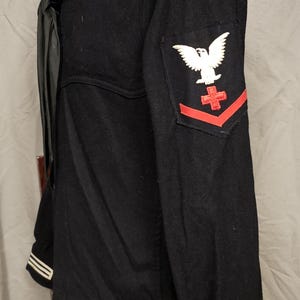 May include: A vintage navy blue sailor's uniform jacket. The jacket features a white collar with three stripes, a black necktie, and a patch with a white eagle and red cross. The sleeves have white stripes.