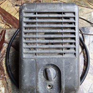 May include: A vintage, gray metal heater with a rectangular shape and horizontal vent slats. The word "Simplex" is visible on the front, along with a circular control knob. A black, circular cord surrounds the heater.