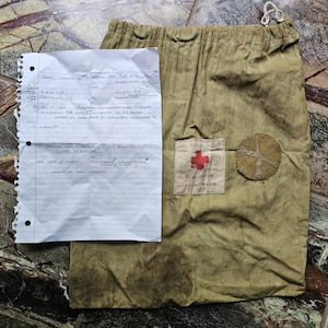 May include: A vintage olive drab canvas bag with a drawstring closure, featuring a Red Cross patch and a circular emblem. A handwritten paper with notes is placed on top of the bag.