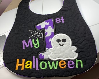 Puede incluir: Babero acolchado negro para bebé con texto bordado morado y verde que dice "My 1st Halloween". Cuenta con un número 1 morado con un fantasma y una aplicación de telaraña. El babero tiene un forro con estampado blanco y morado.
