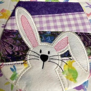 May include: Easter-themed fabric egg with a white bunny applique. The egg is composed of various fabric strips in purple, white, and patterned designs. The bunny has pink inner ears and black whiskers. The background fabric features colorful bunny silhouettes.