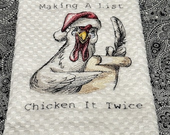 Puede incluir: Toalla de cocina blanca con un bordado de un pollo con un gorro de Papá Noel y sosteniendo un pergamino y una pluma. El texto dice "Making A List Chicken It Twice". La toalla tiene un patrón texturizado.