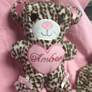 May include: A plush teddy bear with a leopard print pattern in brown and white. The bear has pink accents on the ears, nose, and a heart-shaped patch on its chest with the name "Amber" embroidered in cursive. Small pink bows adorn the feet.