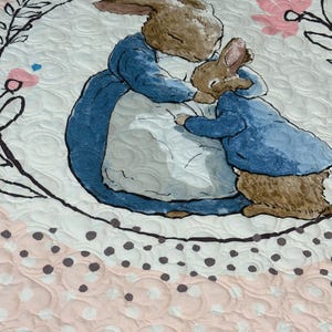 Può includere: Una coperta trapuntata con un'illustrazione di due conigli, uno più grande e uno più piccolo, in un tenero abbraccio. I conigli sono raffigurati in morbidi toni marroni e beige, con abiti blu. Lo sfondo presenta un motivo floreale e a pois.