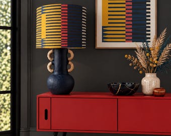 May include: A table lamp with a dark blue textured base featuring two pairs of woven tan handles and a cylindrical lampshade patterned with vertical stripes in yellow, red, and dark blue, sitting on a long red cabinet with black legs.