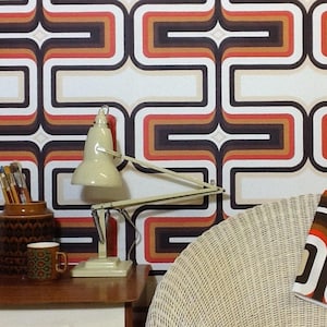 Puede incluir: Una l&aacute;mpara de escritorio de estilo vintage con una pantalla blanca y un brazo negro, sobre un fondo de papel tapiz retro con un patr&oacute;n geom&eacute;trico en tonos marrones, naranjas y rojos.