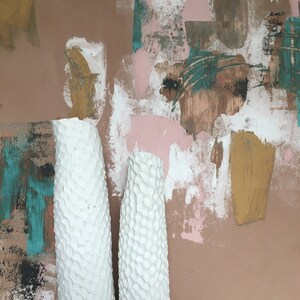 May include: Two white ceramic vases with a textured surface, standing in front of a pink, brown, and teal abstract painted background.