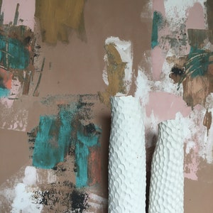May include: Two white ceramic vases with a textured surface. The vases are in front of a wall with abstract painted designs in shades of brown, pink, teal, and white.