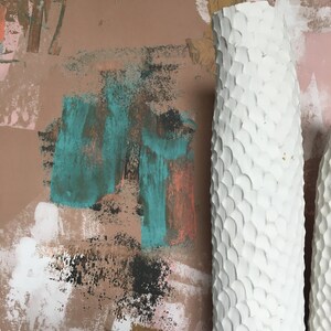 May include: Two white ceramic vases with a textured, scale-like pattern. The vases are against a background of abstract brushstrokes in shades of pink, brown, teal, and gold.