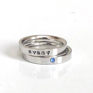 May include: Two silver rings stacked on top of each other. The top ring has the word "evan" engraved on it with a small heart symbol. The bottom ring has a small blue gemstone.