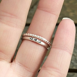 May include: A silver ring with a heart and wings design. The ring is worn on a finger.