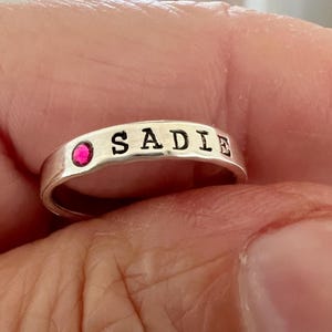 May include: A silver ring with the name "SADIE" stamped on it. The ring has a small, round, pink gemstone on one side.