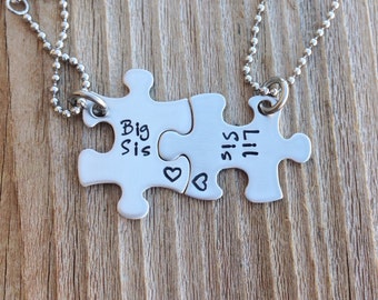 May include: Two silver puzzle pieces on chains, connected by a heart. The pieces say "Big Sis" and "Lil Sis".