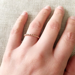 May include: A delicate gold ring with a beaded design. The ring is worn on a finger, showcasing its fine details and subtle shine. The ring's simple yet elegant design makes it a versatile accessory.