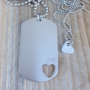 May include: A silver dog tag necklace with a heart cutout and the initials "B&E" engraved on it. The necklace is on a silver chain and is paired with a smaller heart charm with the same initials.
