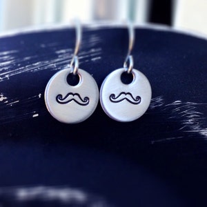 May include: A pair of silver-toned metal drop earrings featuring a circular disc design. Each disc is stamped with a black mustache graphic and hangs from a curved metal ear wire, creating a simple and cohesive set of dangling jewelry pieces.