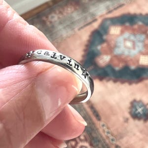 May include: A silver-toned metal ring featuring the name Calvin stamped in lowercase letters on the outer band. The name is flanked by two small, five-pointed star symbols, one on each side of the text, with a smooth, polished finish.