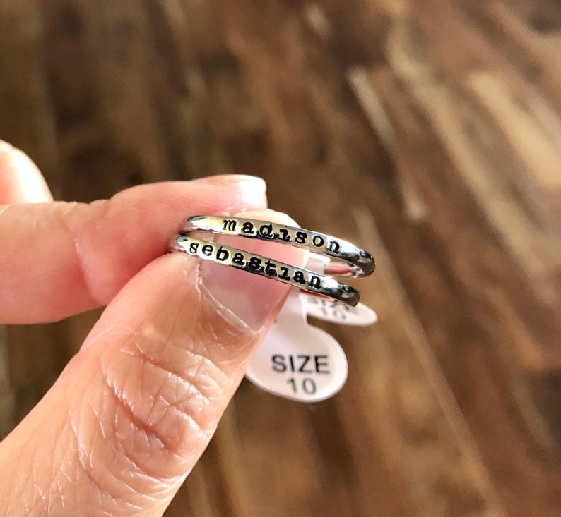 May include: Two silver rings with the names "Madison" and "Sebastian" engraved on them. The rings are stacked on top of each other. A white tag with the text "SIZE 10" is visible in the background.