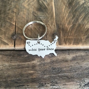 May include: A silver keychain shaped like the United States with a dotted line across the middle and the words "miss your face" inscribed on it. There are two small hearts at the beginning and end of the dotted line.