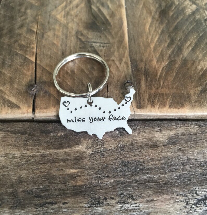 May include: A silver keychain shaped like the United States with a dotted line across the middle and the words "miss your face" inscribed on it. There are two small hearts at the beginning and end of the dotted line.
