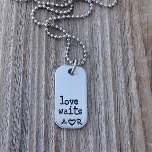 May include: A silver-colored dog tag pendant necklace with a ball chain. The tag has the words "love waits" and the initials "A♡R" stamped in black. The necklace is displayed on a wooden surface.