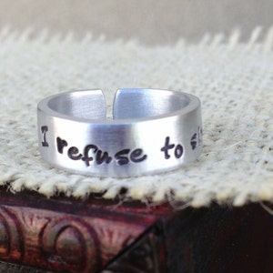May include: A silver-colored adjustable ring with the phrase "I refuse to sink" stamped in black lettering. The ring is made of a thick metal band with an open design. The ring is displayed on a textured, off-white surface.