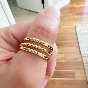 May include: Three gold rings stacked on a finger. The top ring has the word "Maggie" engraved on it. The middle ring is a beaded band. The bottom ring has the word "Hannah" engraved on it.