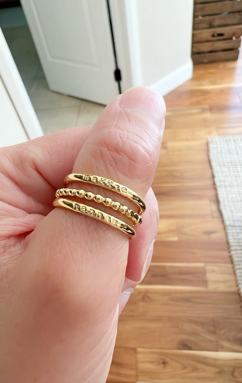 May include: Three gold rings stacked on a finger. The top ring has the word "Maggie" engraved on it. The middle ring is a beaded band. The bottom ring has the word "Hannah" engraved on it.