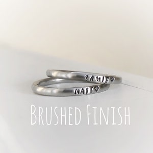 May include: Two brushed metal rings, one stacked on top of the other. The top ring is inscribed with "SAMIE" and a heart symbol, the bottom ring reads "NATE" with a heart. The text "BRUSHED FINISH" is below the rings.