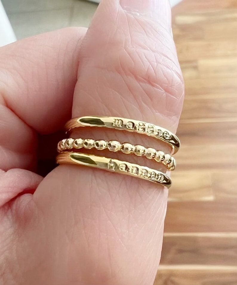May include: Three gold rings stacked on a finger. The top ring has the word "maybe" engraved on it. The middle ring is a beaded band. The bottom ring has the word "tomorrow" engraved on it.
