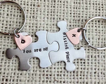 May include: Two silver puzzle piece keychains with a heart charm on each. The heart charms are copper colored and have the letters "D" and "K" engraved on them. The puzzle pieces are engraved with the text "You are my missing piece".