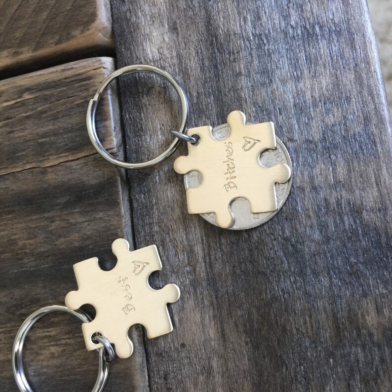 May include: Two gold-toned puzzle piece keychains with a heart symbol and the words "Bitches" and "Best" engraved on them. The keychains are attached to silver key rings.