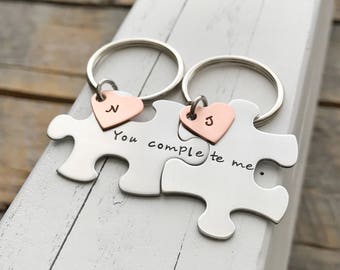May include: Two silver puzzle piece keychains with the words "You complete me." Each keychain has a small, heart-shaped charm with a letter. One charm has the letter "N" and the other has the letter "J".