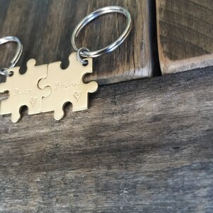 May include: Two gold puzzle pieces attached to a silver key ring. The pieces are engraved with the words "Best" and "Bitches" and a small heart symbol.
