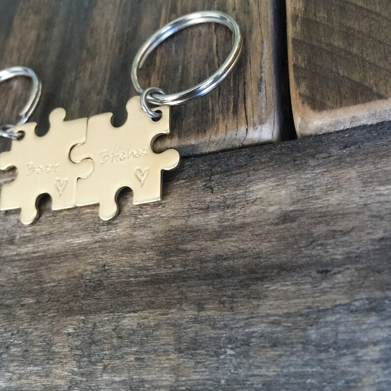 May include: Two gold puzzle pieces attached to a silver key ring. The pieces are engraved with the words "Best" and "Bitches" and a small heart symbol.