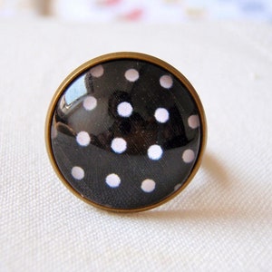 Puede incluir: Un anillo negro con lunares blancos. El anillo es de metal y tiene una parte superior redonda y plana.