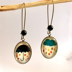 Può includere: Un paio di orecchini pendenti con finitura bronzo. Un orecchino presenta un ombrello nero con cuori rossi, e l'altro orecchino presenta una nuvola blu con gocce di pioggia colorate.