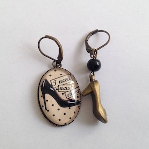 Op de afbeelding: Een paar oorbellen met een zwart-witte schoen met de tekst "Ik heb nieuwe schoenen nodig" en een gouden schoenbedel.