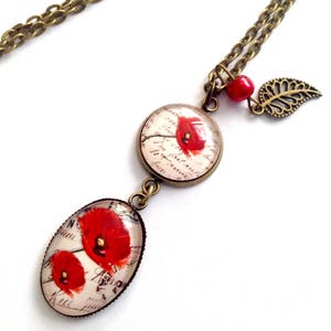 May include: A bronze chain necklace with two glass pendants featuring red poppies on a white background with handwritten text. The necklace also has a small bronze leaf charm and a red bead.