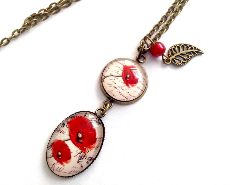 May include: A bronze chain necklace with two glass pendants featuring red poppies on a white background with handwritten text. The necklace also has a small bronze leaf charm and a red bead.