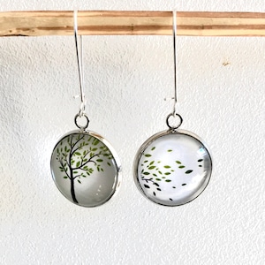 Peut inclure: Une paire de boucles d'oreilles en argent avec des cabochons ronds en verre. Les cabochons présentent un arbre vert avec des feuilles qui soufflent dans le vent. Une boucle d'oreille montre l'arbre avec des feuilles, l'autre montre l'arbre avec des feuilles qui s'envolent.