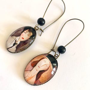 Könnte beinhalten: Ein Paar ovale Ohrringe mit einem bronzefarbenen Metallrahmen. Jeder Ohrring hat einen Glas-Cameo mit einem Gemälde eines Frauen Gesichts. Die Ohrringe sind mit einer schwarzen Perle verziert.