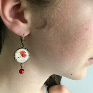 May include: A pair of dangle earrings with a red poppy flower design inside a clear glass dome. The earrings are made of bronze and have a small red bead hanging from the bottom.