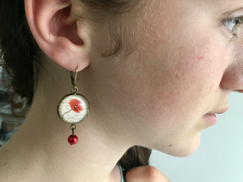 May include: A pair of dangle earrings with a red poppy flower design inside a clear glass dome. The earrings are made of bronze and have a small red bead hanging from the bottom.