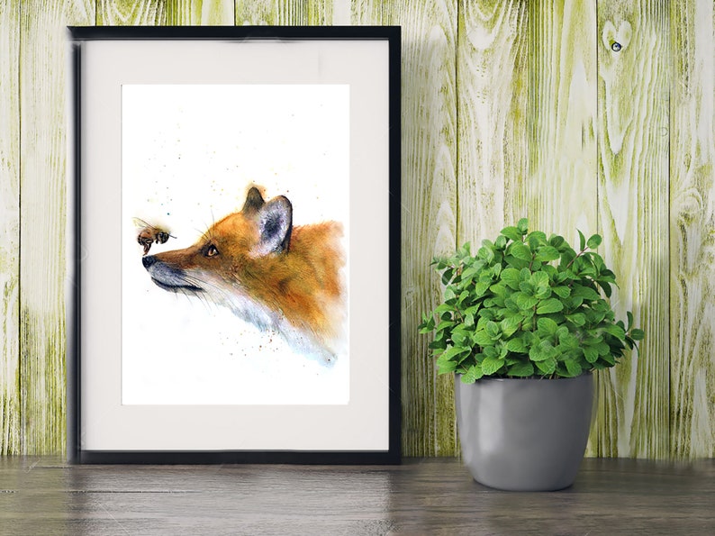 May include: A watercolour painting of a fox's head with a bee flying in front of it. The fox is looking at the bee with a curious expression. The painting is framed in a black frame and is set against a wooden background.