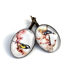Peut inclure: Une paire de boucles d'oreilles ovales avec une finition bronze. Chaque boucle d'oreille présente un cabochon en verre transparent avec une image peinte d'un oiseau perché sur une branche avec des fleurs roses.