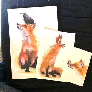 May include: Three watercolour paintings of foxes. The first painting features a fox with a black crow perched on its head. The second painting features a fox with a squirrel perched on its head. The third painting features a fox looking at a bee.