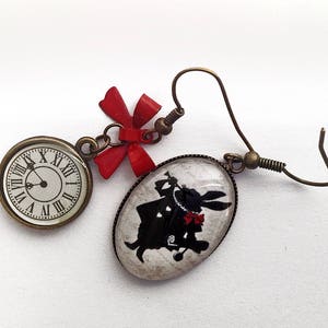 Peut inclure: Une paire de boucles d'oreilles avec un charme de nœud rouge, un charme de cadran d'horloge et un charme en verre avec une silhouette du Lapin Blanc d'Alice au Pays des Merveilles.
