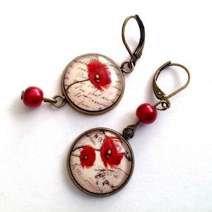 May include: A pair of earrings with red poppy flower designs on a white background with a handwritten script. The earrings are made with bronze-colored metal and red glass beads.