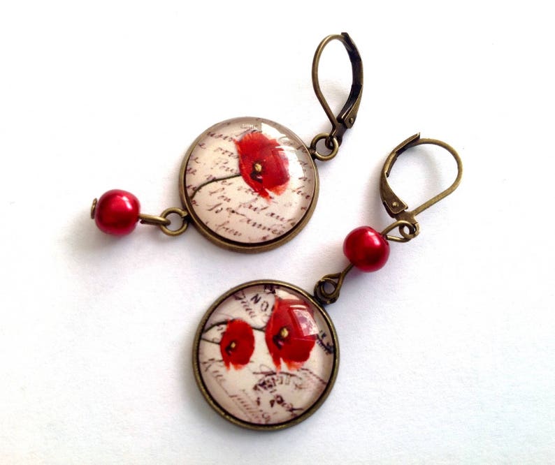 May include: A pair of earrings with red poppy flower designs on a white background with a handwritten script. The earrings are made with bronze-colored metal and red glass beads.